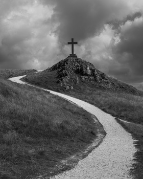 A winding path leads up a hillside toward a cross silhouetted against a dramatic cloudy sky.