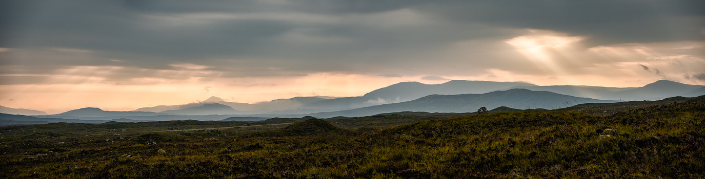 Vast moorland landscape with distant mountains beneath dramatic cloudy sky with golden light breaking through.