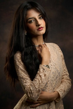 Woman with long dark hair wearing an ornate beige embroidered top against a dark brown background.