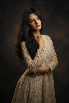 Woman with long dark hair wearing a beige embroidered dress against a dark brown backdrop.