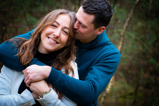 Man in blue jacket embracing smiling woman outdoors with blurred green foliage background.
