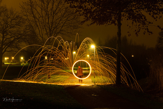 Light painting photography with spinning steel wool creating golden arcs against a dark wooded backdrop.