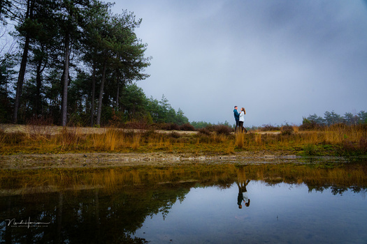Two people standing on grassy wetland bank beside still water reflecting sky and trees.