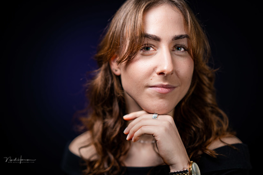 Woman with long wavy brown hair gazing thoughtfully to the side against a dark background.