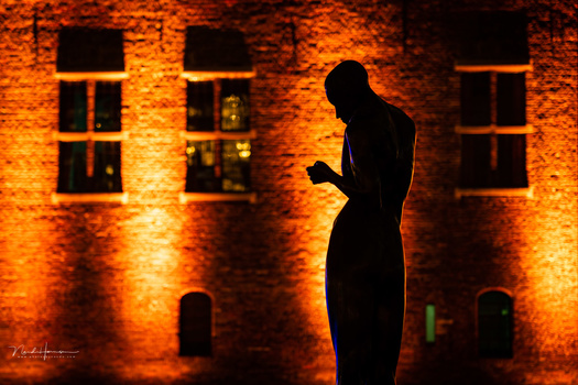 Silhouetted figure standing against illuminated brick building facade with windows at night.