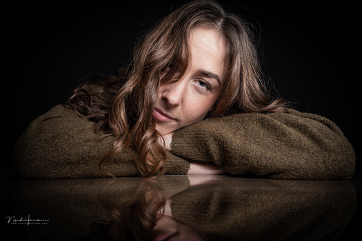Woman with long wavy brown hair resting her head on her arms against a reflective dark surface.