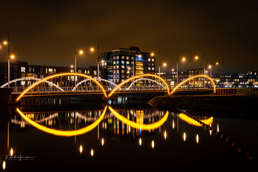 Illuminated arched bridge reflecting in still water at night with city buildings in background.