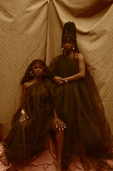 Two figures in flowing black tulle dresses posed against draped tan fabric backdrop.