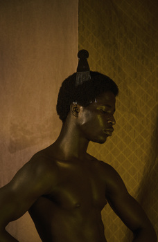 Shirtless male figure with dark skin tone and braided hair, photographed in profile against a textured wall with warm lighting.