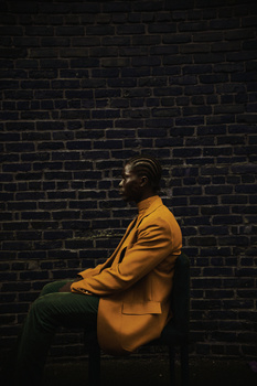 Man in mustard yellow jacket seated against dark brick wall in profile.