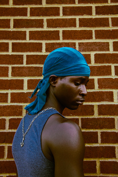 Person in blue du-rag and sleeveless top posed against a brick wall.