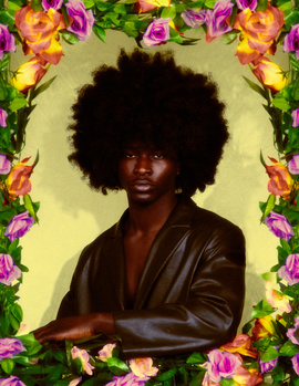 Portrait of a person with a natural afro hairstyle wearing a brown leather jacket against a yellow background surrounded by vibrant flowers.