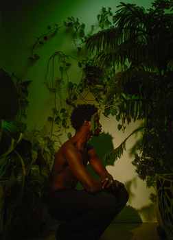 Shirtless man crouching against green-lit wall with hanging plants and foliage.