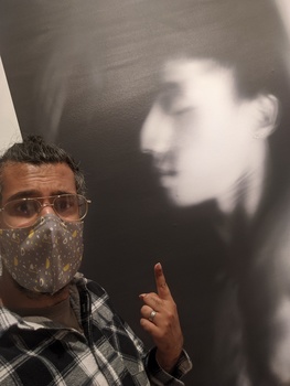 Man wearing glasses and plaid shirt pointing at black and white portrait projected or displayed on wall behind him.