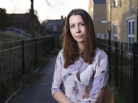 Woman with long dark hair wearing a patterned lavender shirt, standing on a pathway between black metal railings with stone buildings in the background.