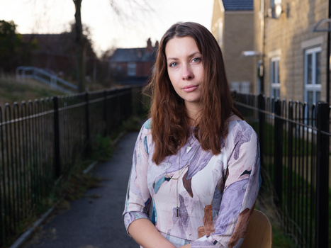 Woman with long brown hair wearing a patterned lavender shirt standing on a tree-lined path.