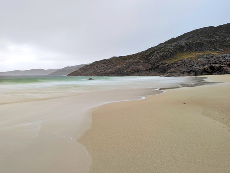 Sandy beach with turquoise shallow water and dark rocky hillside under overcast sky.