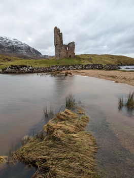 Ruined stone tower on a hillside reflected in a shallow tidal inlet with grassy foreground.