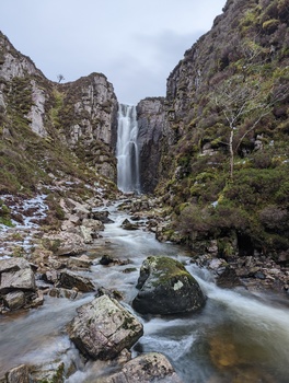 Waterfall cascading between steep moss-covered rock cliffs with a flowing stream in the foreground.