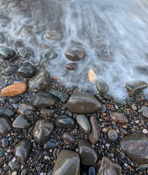 Smooth rocks and pebbles on a beach shore with shallow water and foam washing over them.