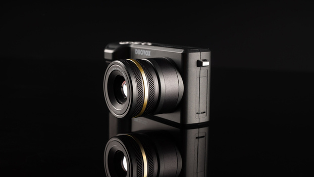 Compact camera with prime lens and gold accents reflected on black surface.