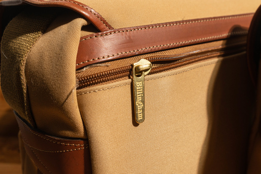 Close-up of a tan canvas and brown leather bag with brass zipper hardware and stitched details.