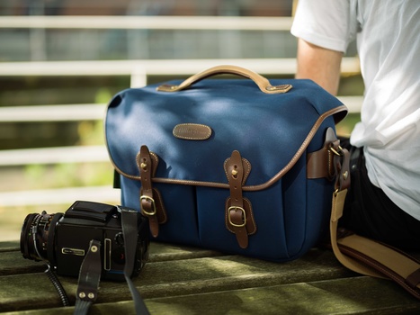 Blue leather camera bag with brown leather accents and brass hardware sits on a wooden bench next to a smaller black camera bag.