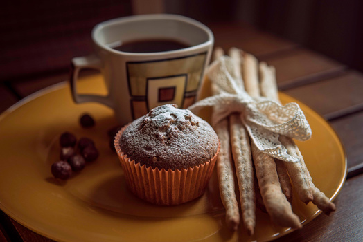 Muffin dusted with powdered sugar on a yellow plate next to breadsticks and a coffee cup.