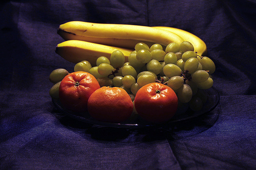 Still life arrangement of bananas, green grapes, and red tomatoes on a black plate against deep purple fabric.
