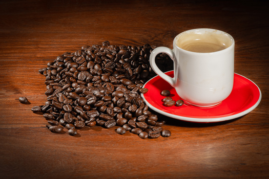 White ceramic mug filled with coffee on a red saucer, positioned next to scattered dark roasted coffee beans on a wooden surface.