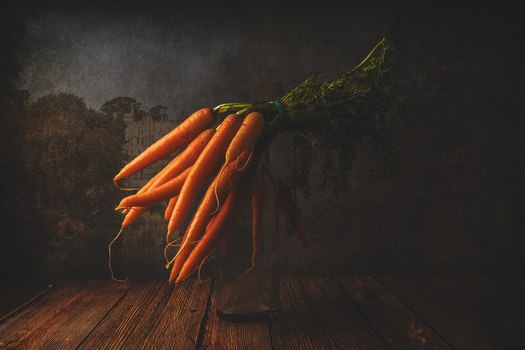 Bundle of fresh carrots with green tops on weathered wooden surface against dark background.