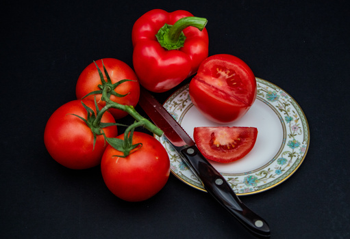 Fresh red tomatoes and bell pepper arranged with a decorative plate and knife on dark background.
