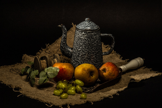Still life arrangement with ornate metal teapot, fresh fruits, and vintage objects on burlap against black background.