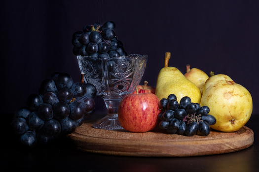 Still life arrangement of dark grapes, pears, pomegranate, and a crystal glass on wooden board against black background.