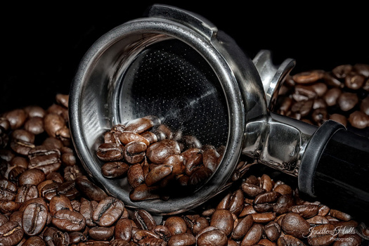 Espresso portafilter filled with roasted coffee beans surrounded by scattered beans on black background.