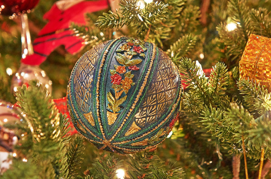Ornate blue and gold beaded Christmas ornament hanging on evergreen tree branch among lights and red poinsettia.