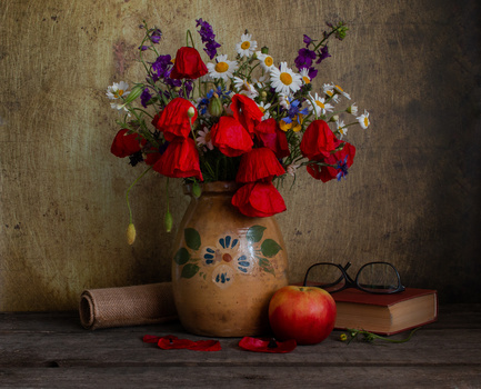 Still life arrangement featuring red poppies and daisies in a decorated ceramic vase beside books, glasses, and an apple.