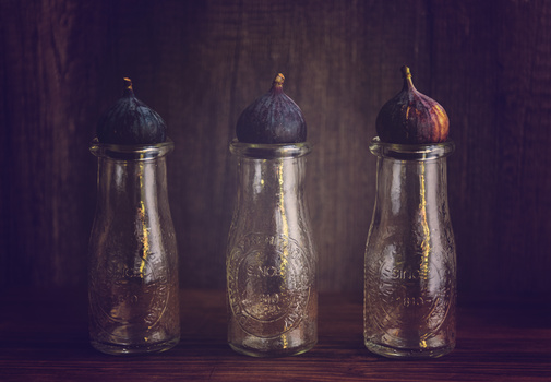 Three glass bottles with dark figs on top arranged in a row against a textured brown background.