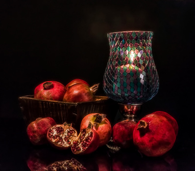 Still life arrangement of pomegranates with a decorative glass vase against black background.