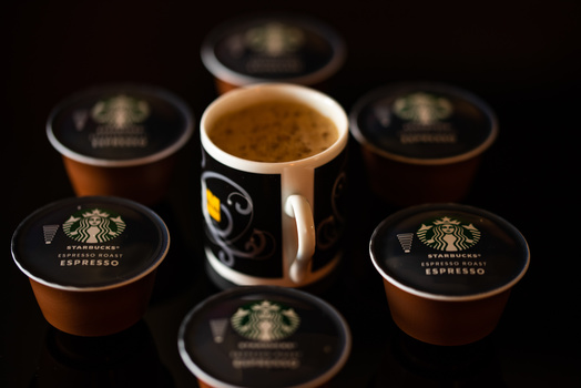 Six espresso capsules arranged in a circle around a center cup filled with dark brown espresso on black background.