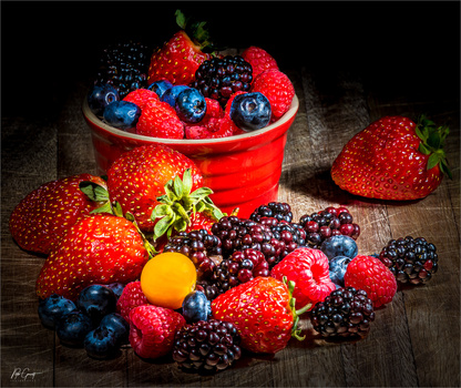 Assorted fresh berries arranged on wooden surface with red cup in background.