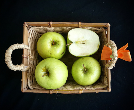 Four green apples and one halved apple arranged in a woven basket with rope handles against a black background.