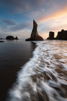 Dramatic sea stack rising from ocean during golden hour with foaming waves in foreground.