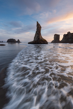 Dramatic sea stacks rising from shallow water at sunset, with foaming waves in the foreground.