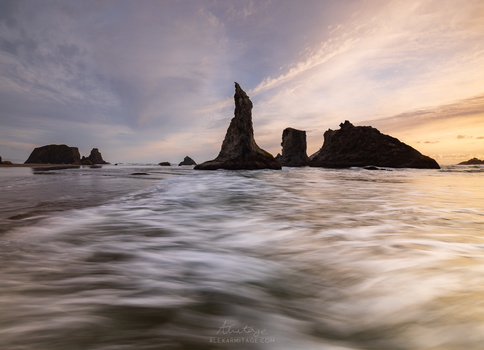Dramatic sea stacks rising from shallow water at sunset with motion-blurred waves in foreground.