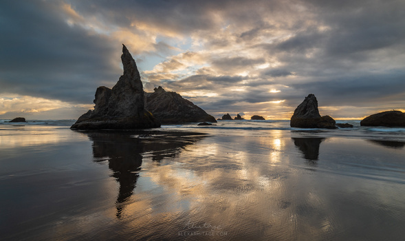 Dramatic rock formations reflected in wet sand at sunset with stormy clouds overhead.