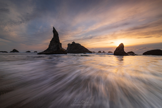 Silhouetted sea stacks rising from flowing water at sunset with dramatic cloudy sky.