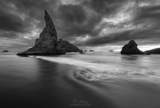 Dramatic black and white seascape with a tall rocky pinnacle emerging from the ocean and stormy clouds above.