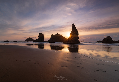 Dramatic coastal rock formations silhouetted against a golden sunset on a wide sandy beach.