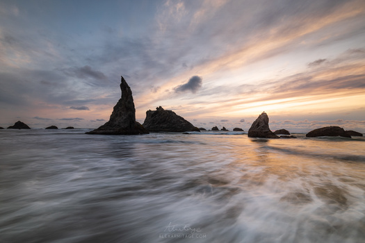 Dramatic rocky sea stacks silhouetted against a golden sunset sky with soft waves washing across the foreground beach.